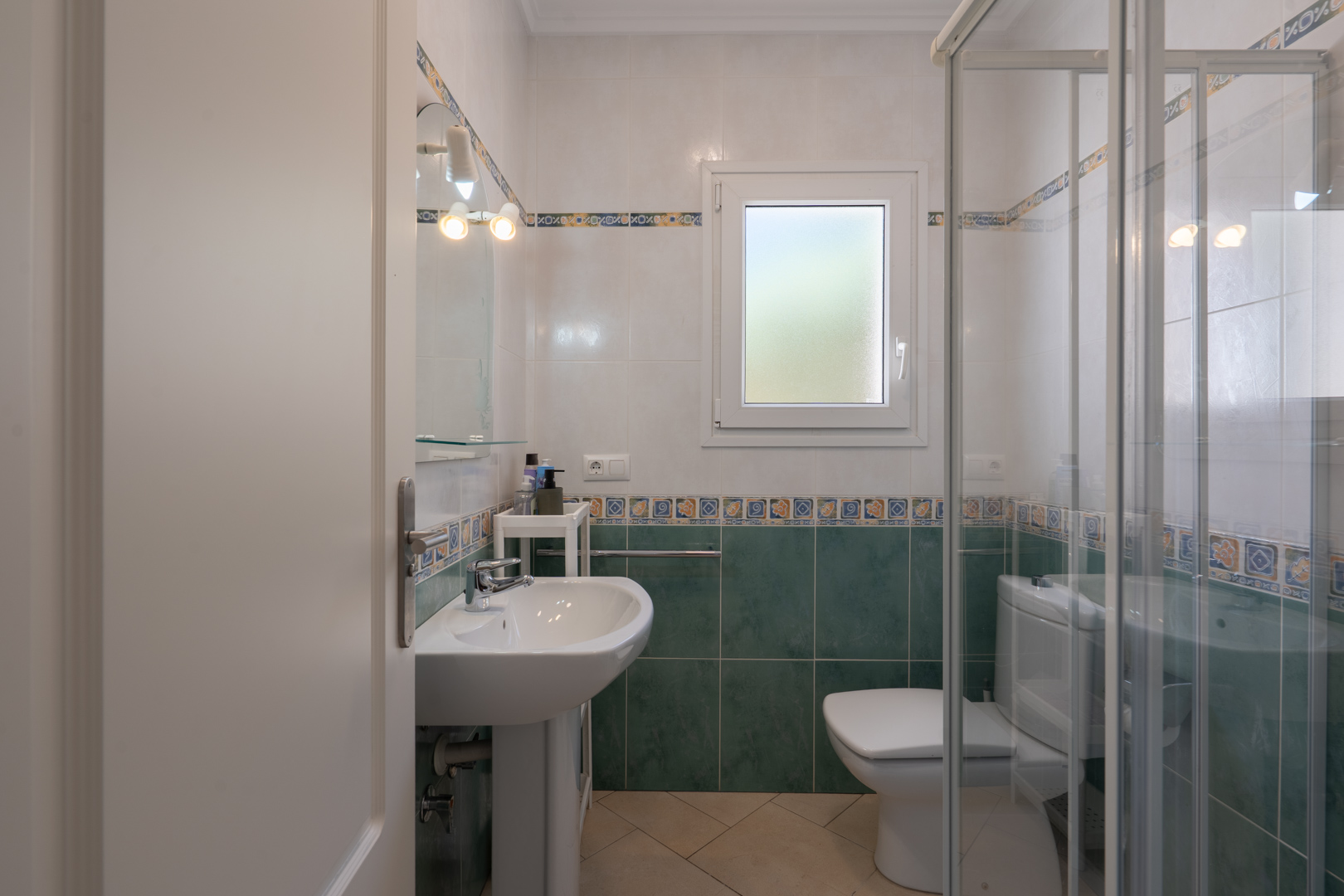 Functional bathroom featuring green and white tiling, a pedestal sink, a glass-enclosed shower, and a frosted PVC window for natural light.