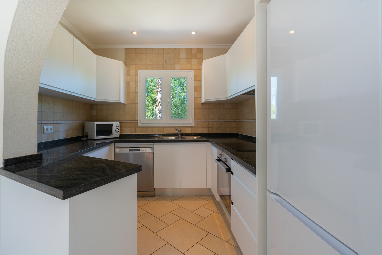 Functional U-shaped kitchen featuring dark granite countertops, white cabinetry, integrated appliances, and a PVC window overlooking the garden.