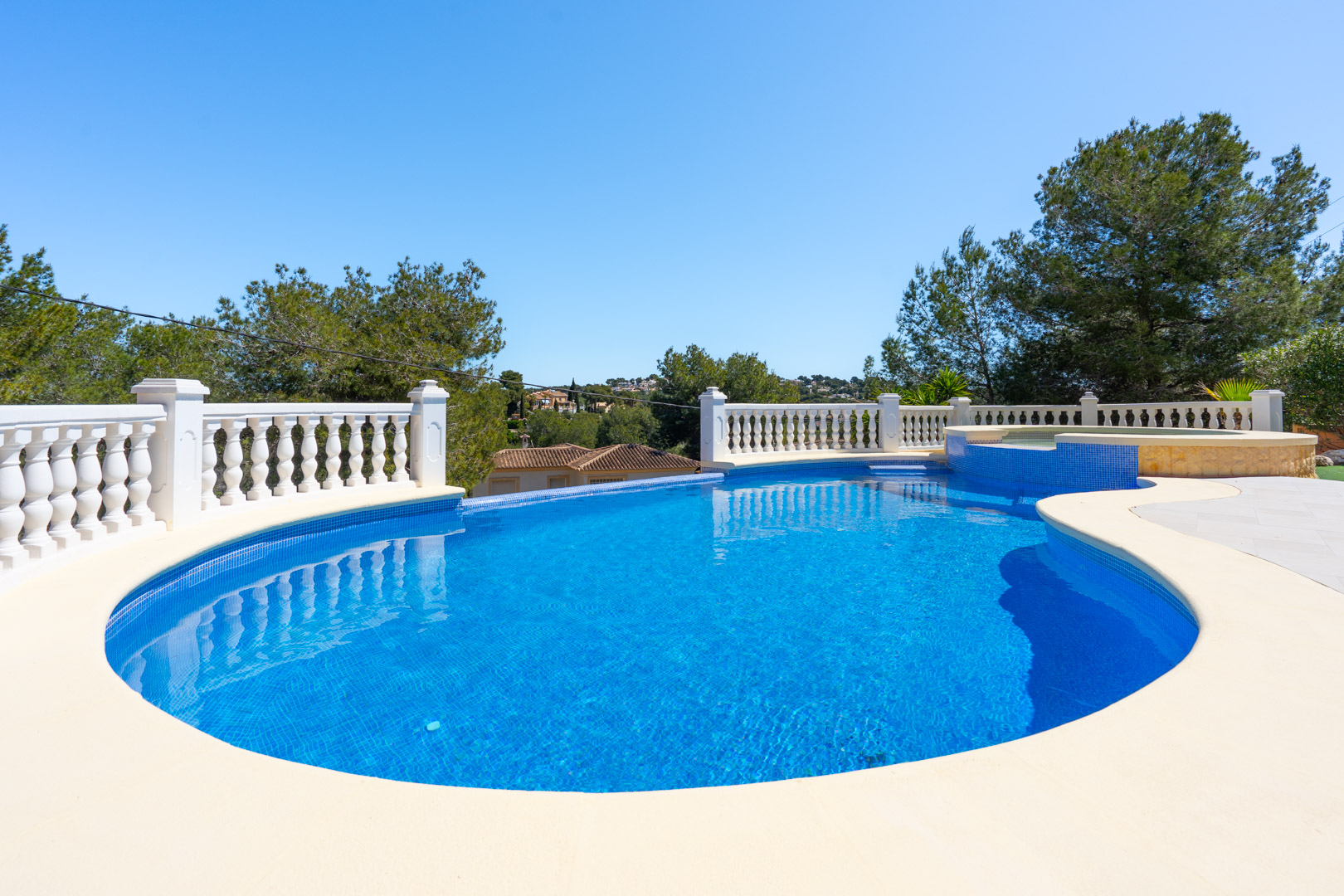 Irregularly shaped outdoor pool featuring blue mosaic tiling, a classic white balustrade, and a sun terrace overlooking the Mediterranean pine landscape.