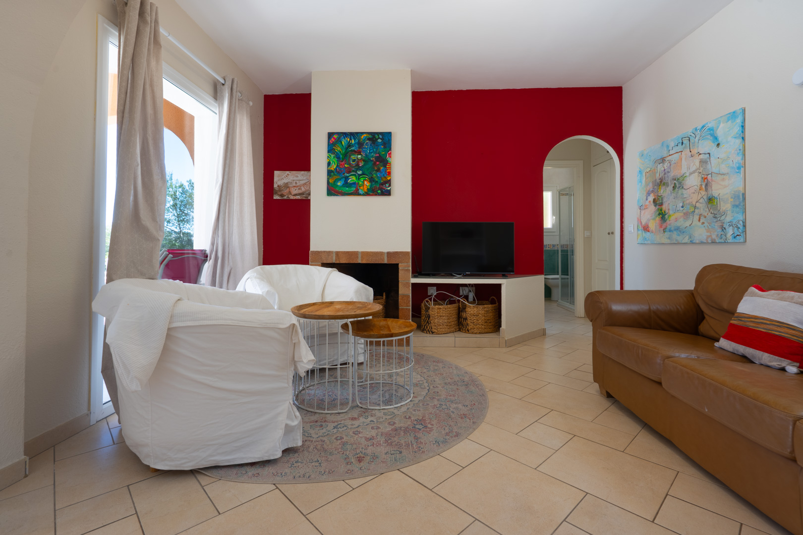 Living area featuring a red accent wall, built-in fireplace, and arched doorway. The space includes tiled flooring, leather seating, and large windows.