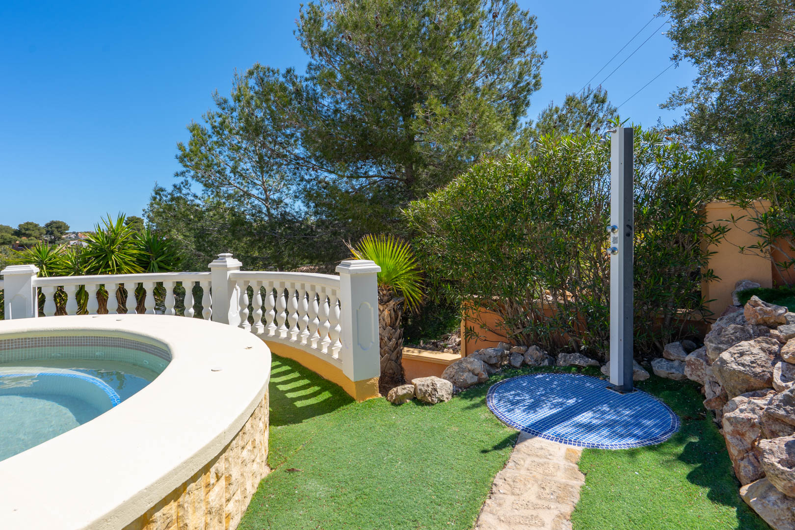 Outdoor shower with a blue mosaic tile base, positioned next to the curved pool edge and framed by a white balustrade and Mediterranean trees.