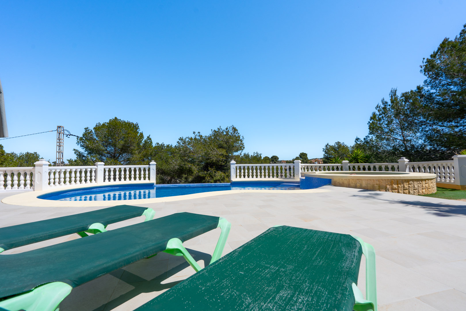 Outdoor terrace featuring a blue-tiled swimming pool with a white balustrade, integrated stone jacuzzi, and sun loungers set against a backdrop of Mediterranean pines.