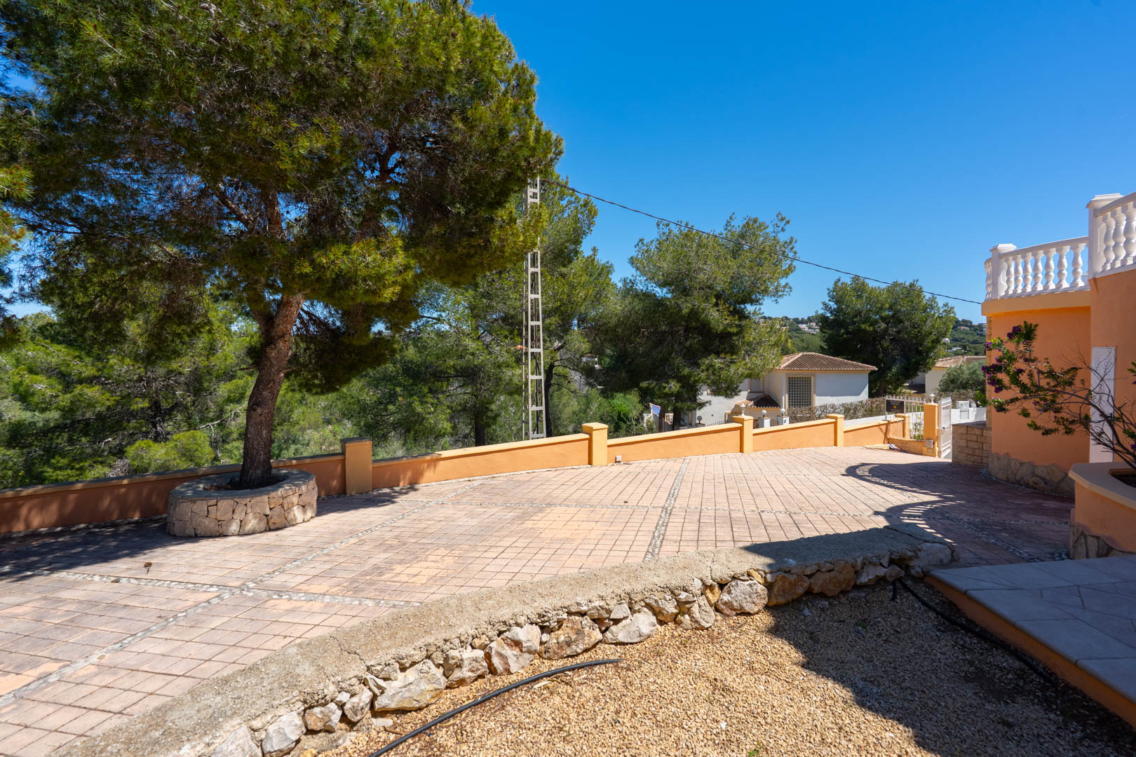 Spacious paved entrance area featuring patterned tiling, a perimeter wall, and a mature pine tree set within a natural stone circular planter.