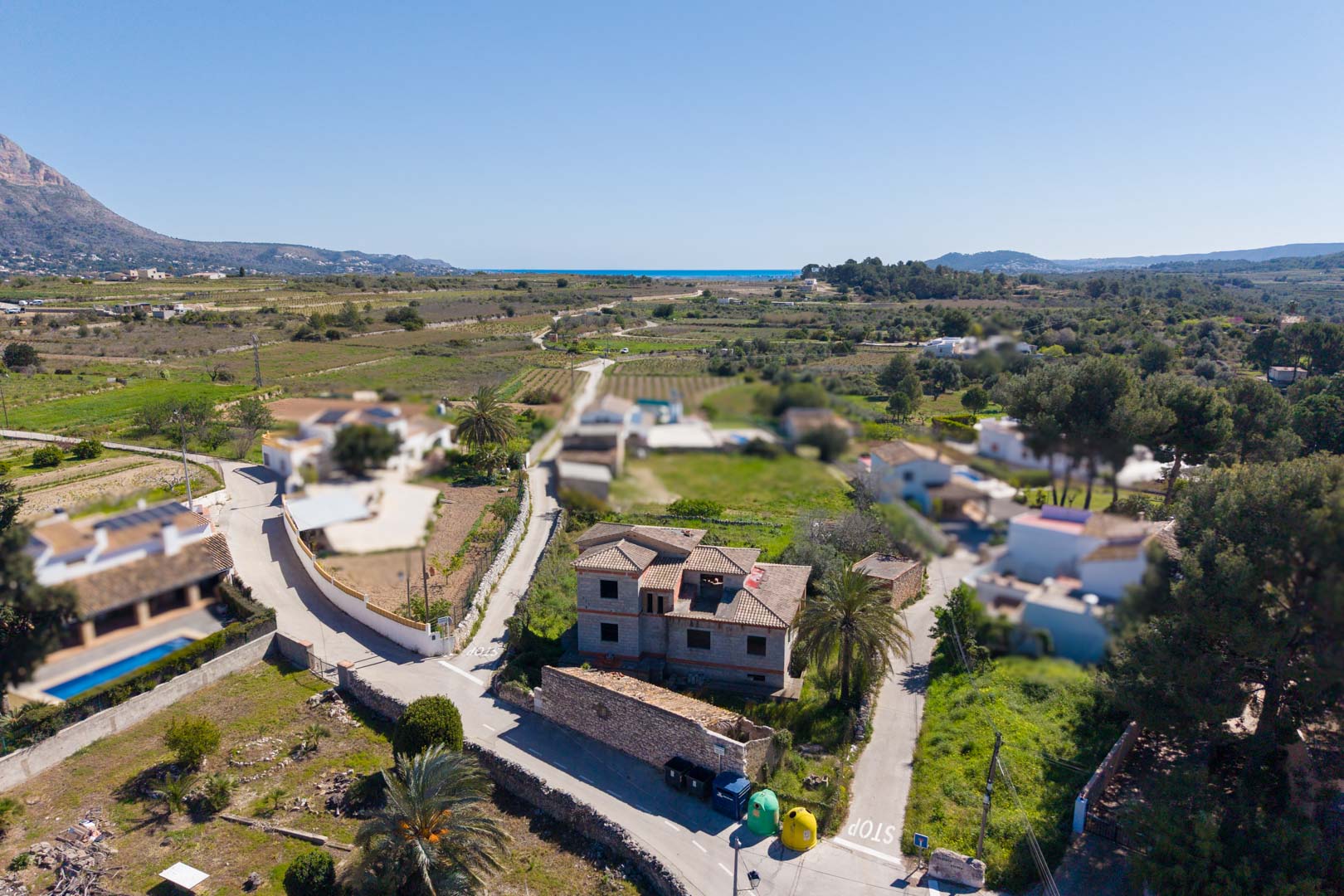 Aerial view showing the unfinished house structure with a tiled roof, set within a rural landscape with views of Montgó mountain and the distant sea.