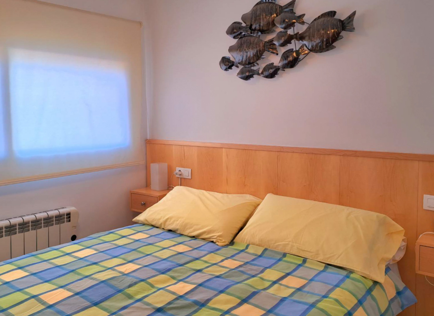 Bedroom featuring an integrated wooden headboard with matching nightstands and metal wall art. Equipped with an electric radiator and a window with a roller blind.