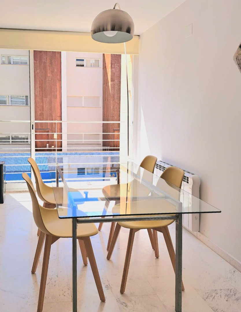 Bright dining area featuring a glass table, Nordic-style chairs, and marble flooring, offering views of the communal pool through large sliding doors.