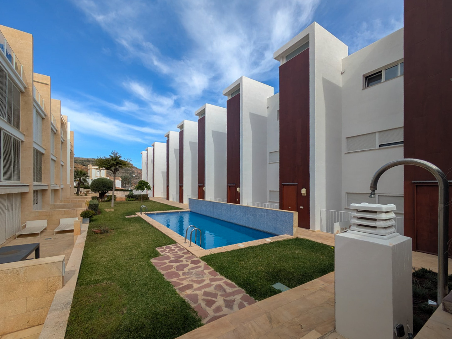 Communal area featuring a rectangular swimming pool with blue mosaic tiles, manicured lawns, and stone paths set against modern white and terracotta buildings.