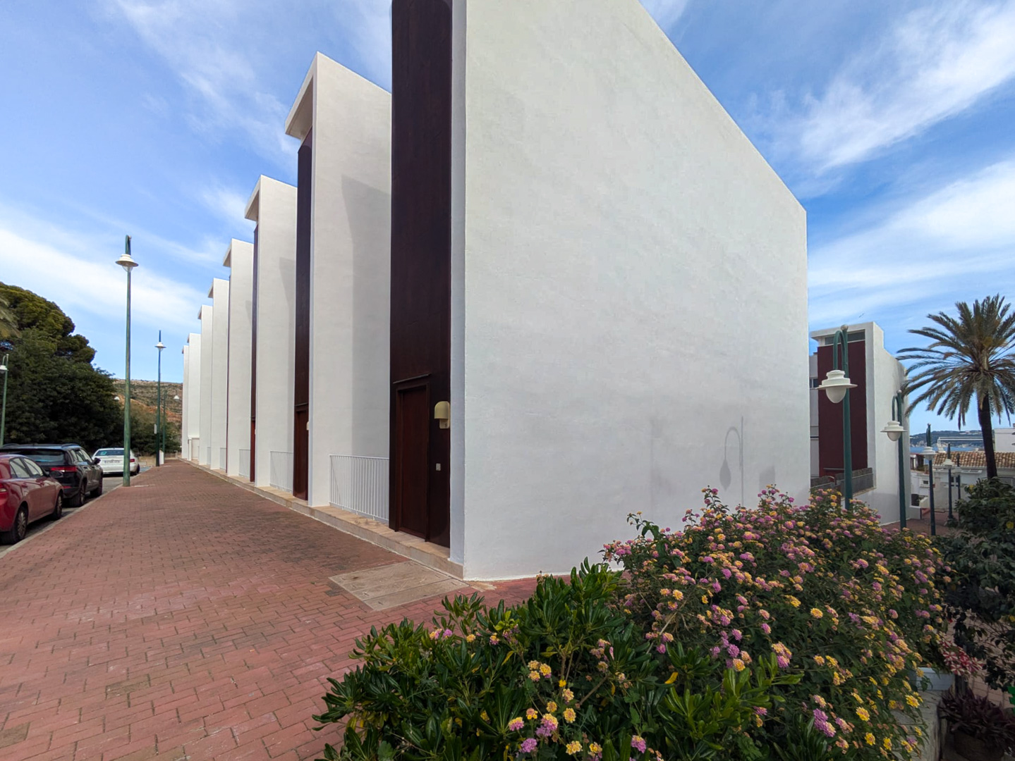 Contemporary exterior design featuring a minimalist white facade with dark vertical accents, set along a paved walkway with flowering Mediterranean shrubs.