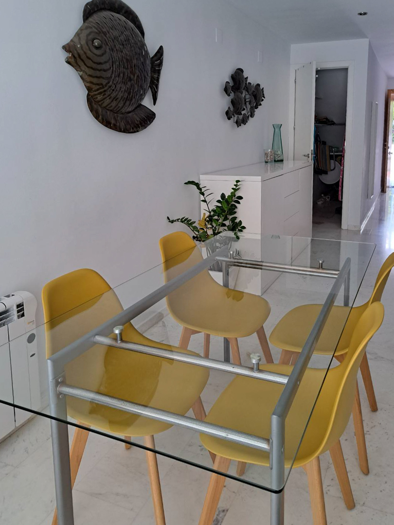 Dining area featuring a glass-top table with yellow chairs and marble flooring. The space includes a white sideboard, electric radiator, and marine-themed wall art.