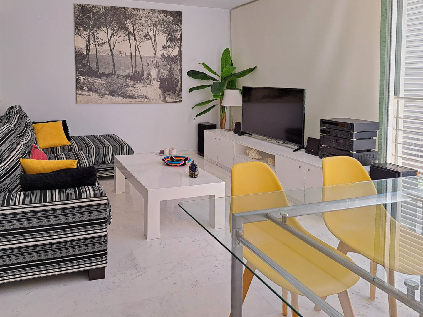 Living-dining room featuring marble floors, a striped sofa, and a glass table with yellow chairs. A white media console and large wall art define the space.