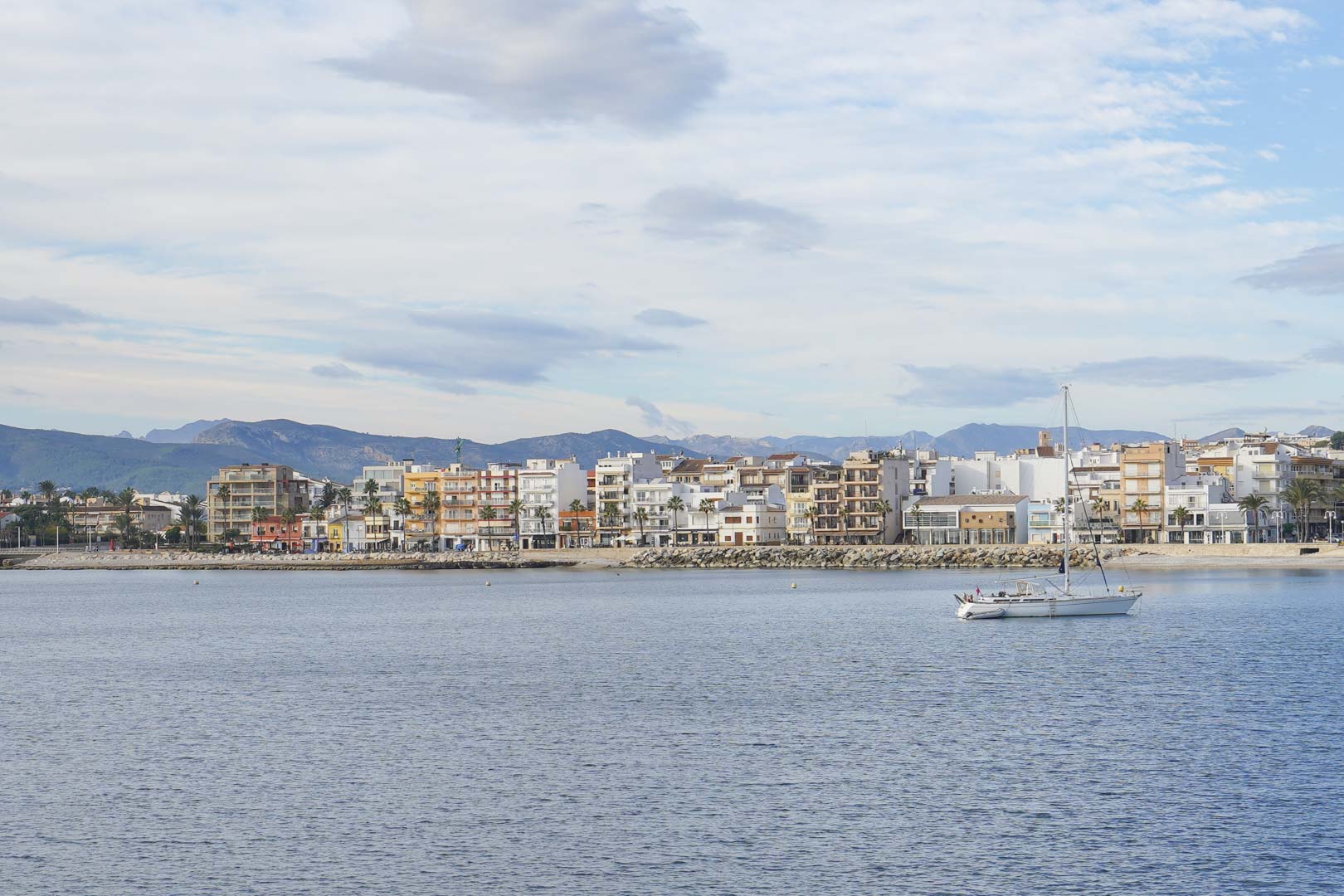 Panoramic view of the Jávea port coastline from the water, featuring seafront buildings, a sailboat, and the mountain range in the background under a soft sky.