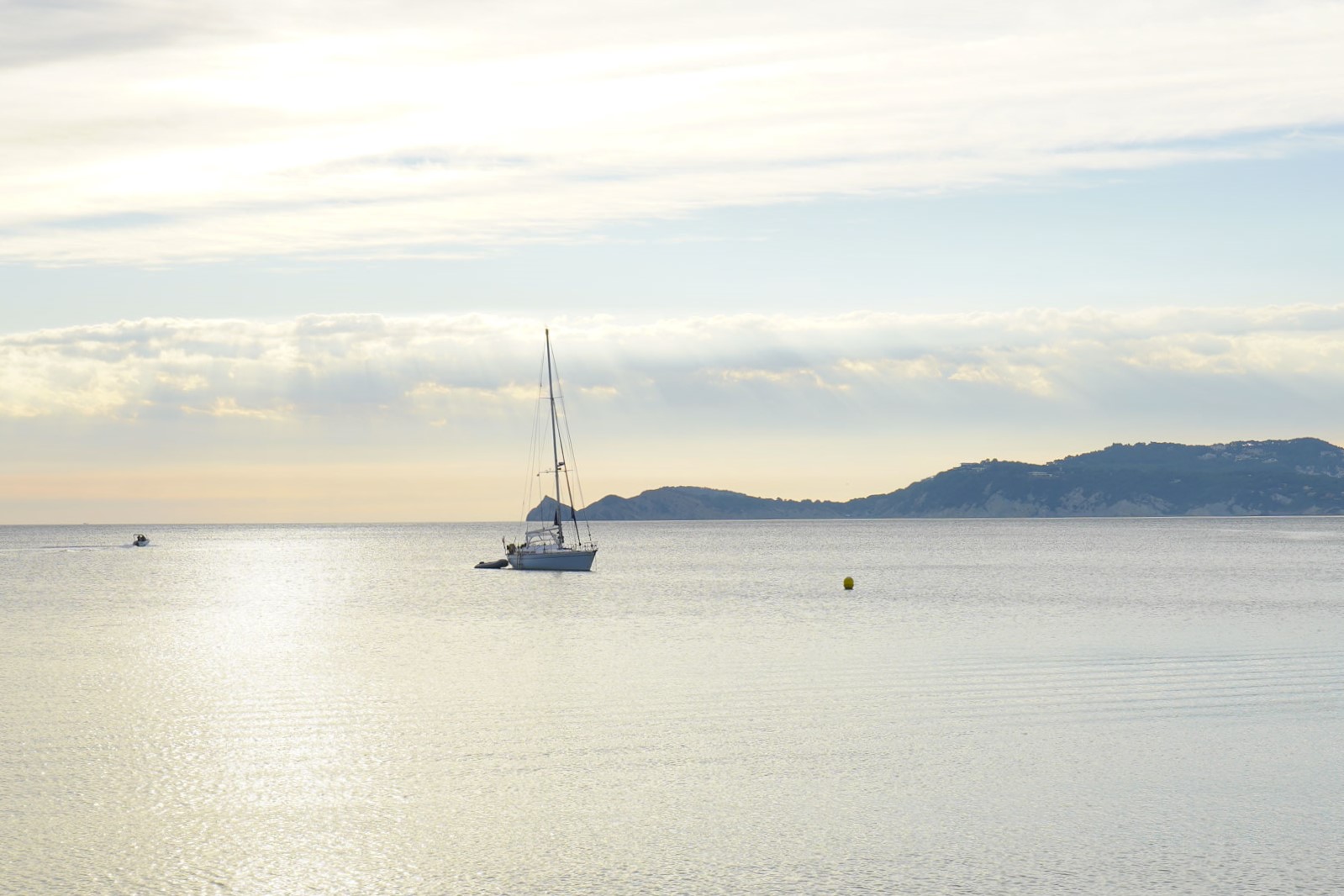 View of the Mediterranean Sea near Jávea port, featuring a sailboat on calm waters with soft natural light and the coastal silhouette in the background.