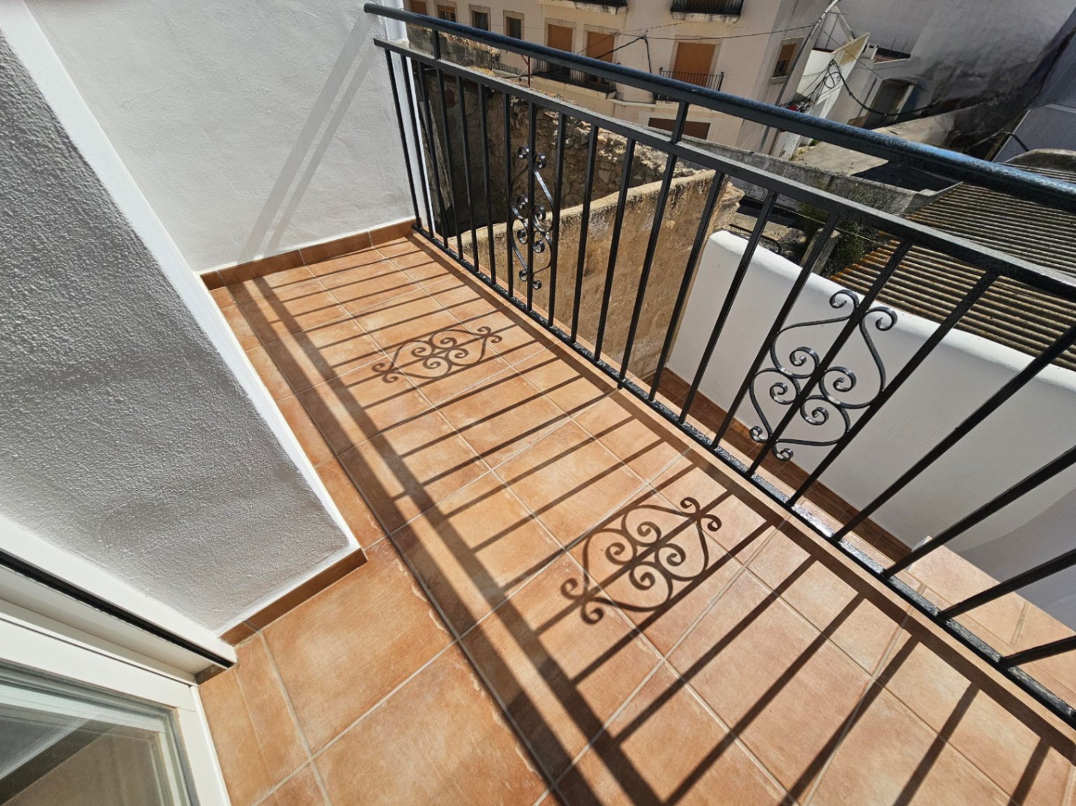 Balcony featuring terracotta floor tiles and a black wrought-iron railing with decorative scrollwork, overlooking the traditional village rooftops.
