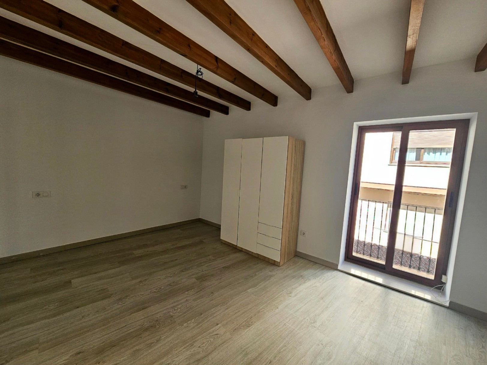 Bedroom featuring exposed wooden ceiling beams, light laminate flooring, and a sliding glass door leading to a private balcony with wrought-iron details.