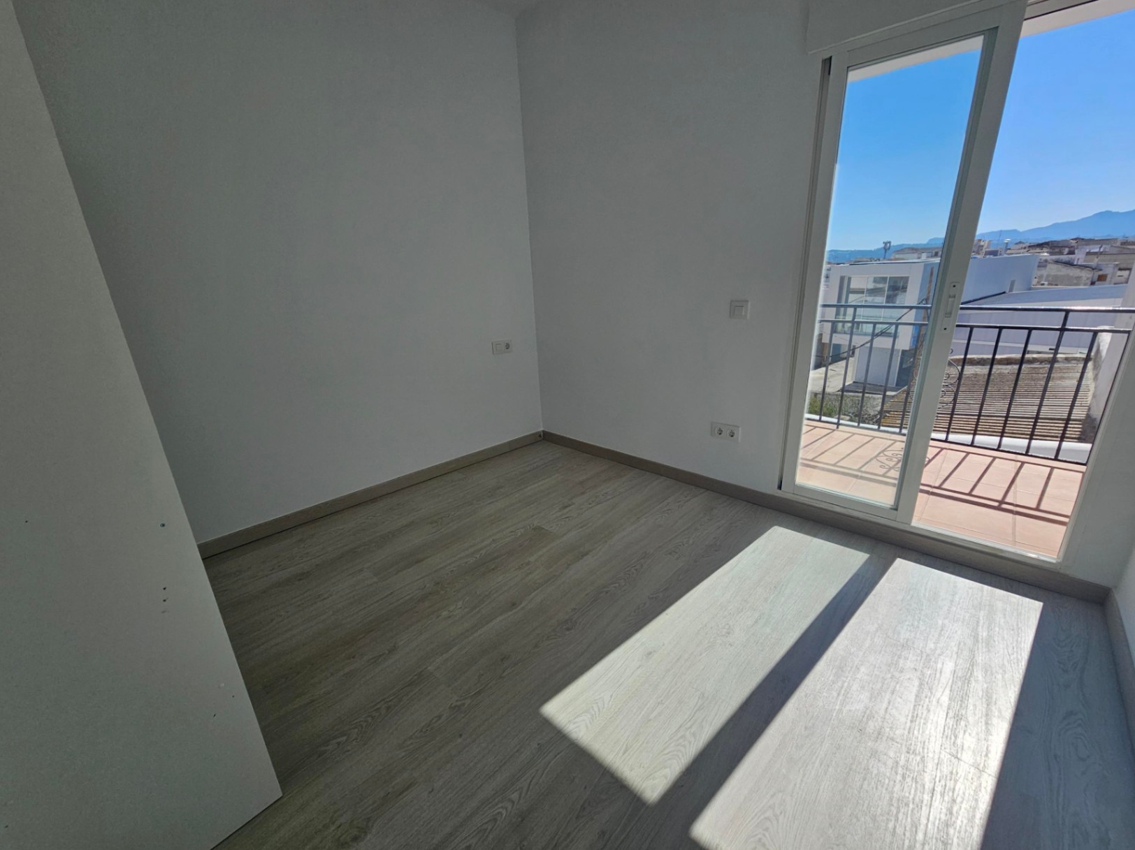 Bright bedroom featuring light wood flooring and direct access to a private balcony with open views of the village and distant mountains.