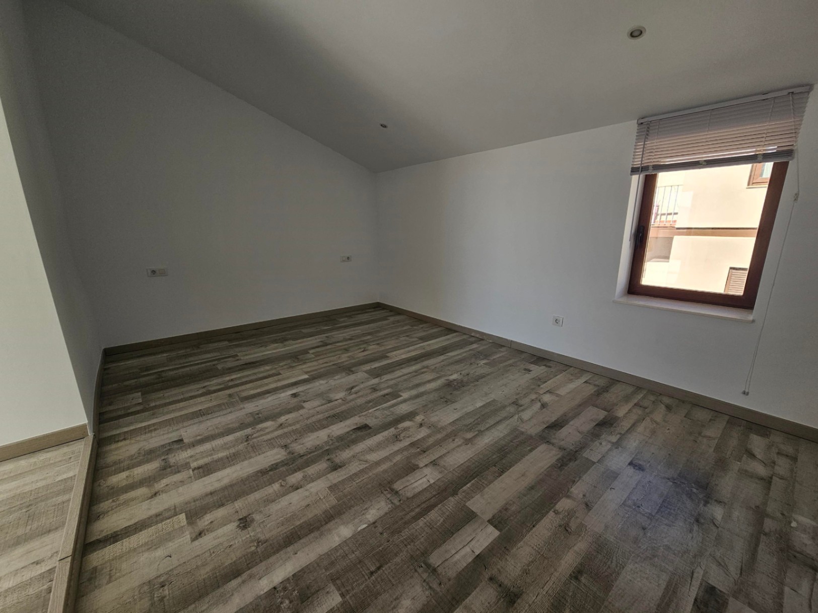 Bright room featuring rustic wood-effect flooring, white walls, and a sloped ceiling with recessed spotlights. A window with blinds provides natural light.
