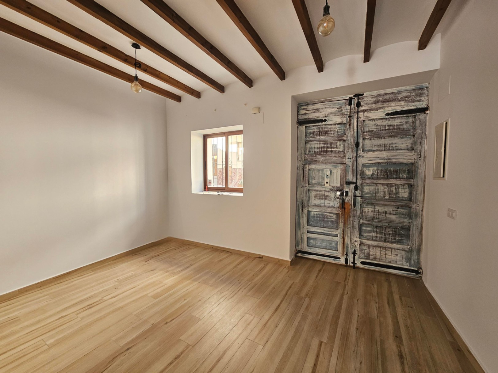 Interior space featuring exposed dark wood ceiling beams, light flooring, and a large rustic double door with a distressed finish and traditional iron hardware.
