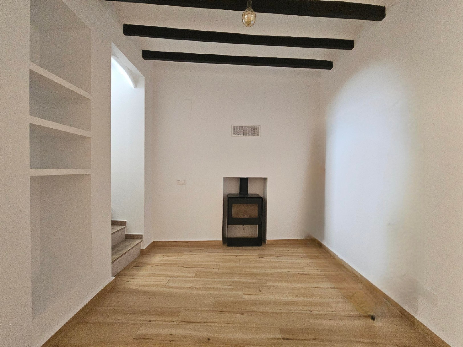 Living area featuring dark wooden ceiling beams, a built-in shelving unit, and a recessed wood burner. Light wood-effect flooring complements the clean white walls.