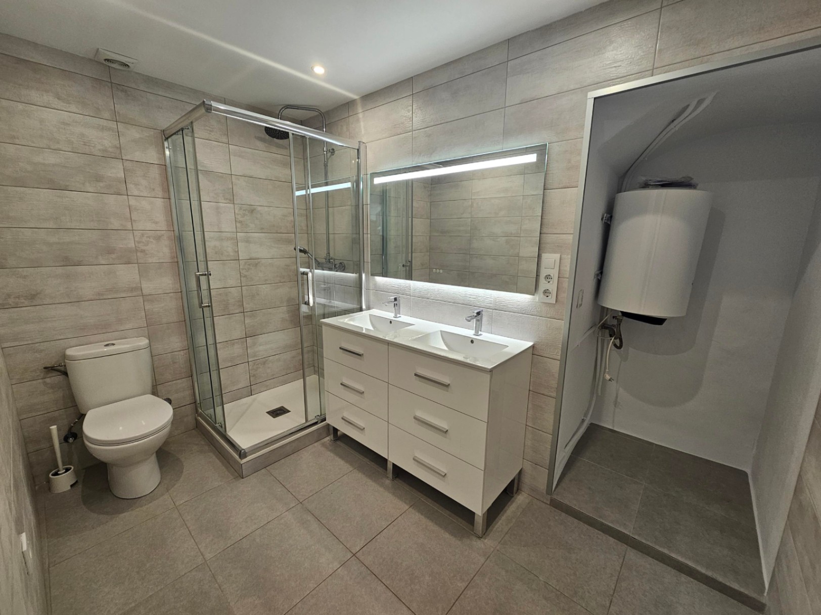 Modern bathroom featuring a glass-enclosed shower, white double vanity with integrated sinks, backlit mirror, and neutral stone-effect wall tiling.