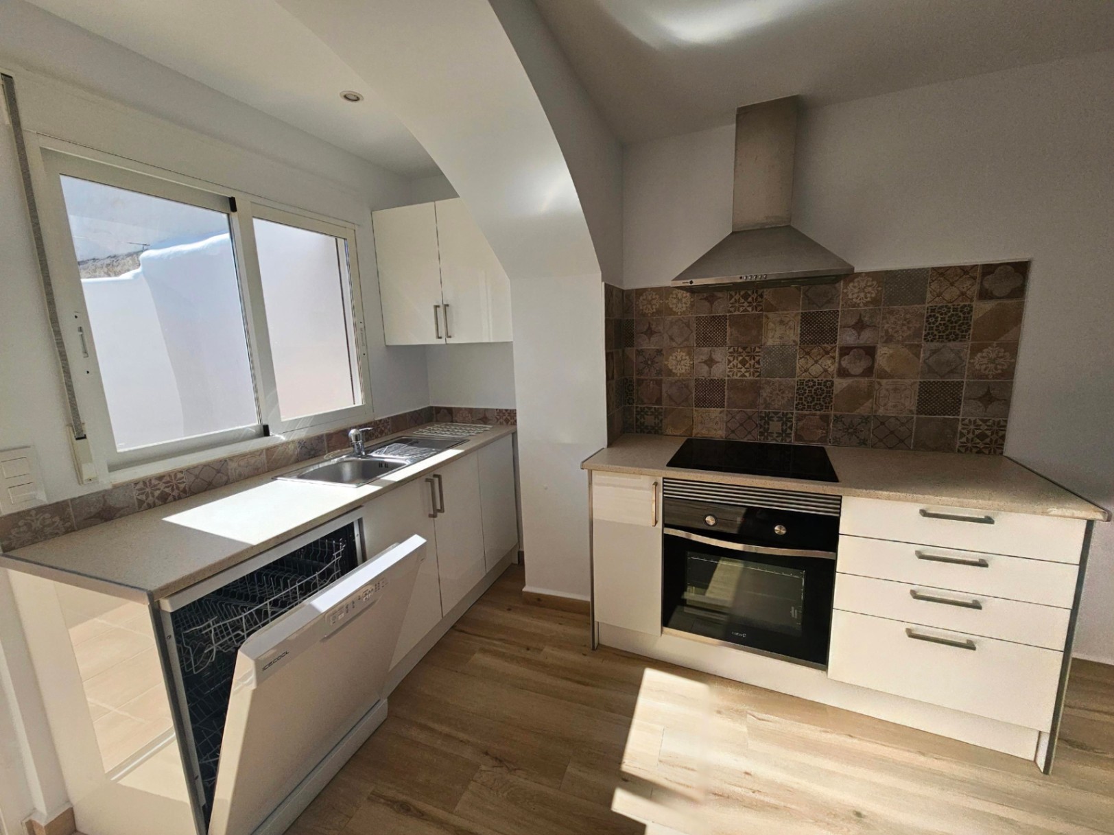 Modern renovated kitchen featuring white cabinetry, patterned tile backsplash, and integrated appliances. Natural light enters through the window above the sink area.