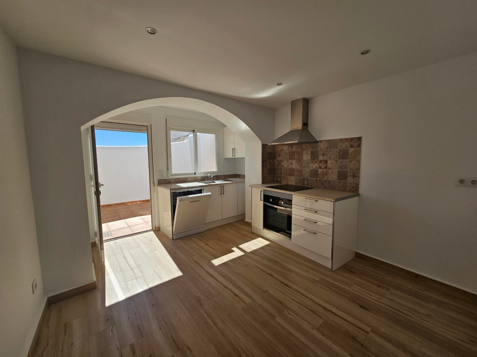 Open-plan kitchen featuring white cabinetry, patterned tile backsplash, and recessed lighting, with an arched doorway leading to the private patio.