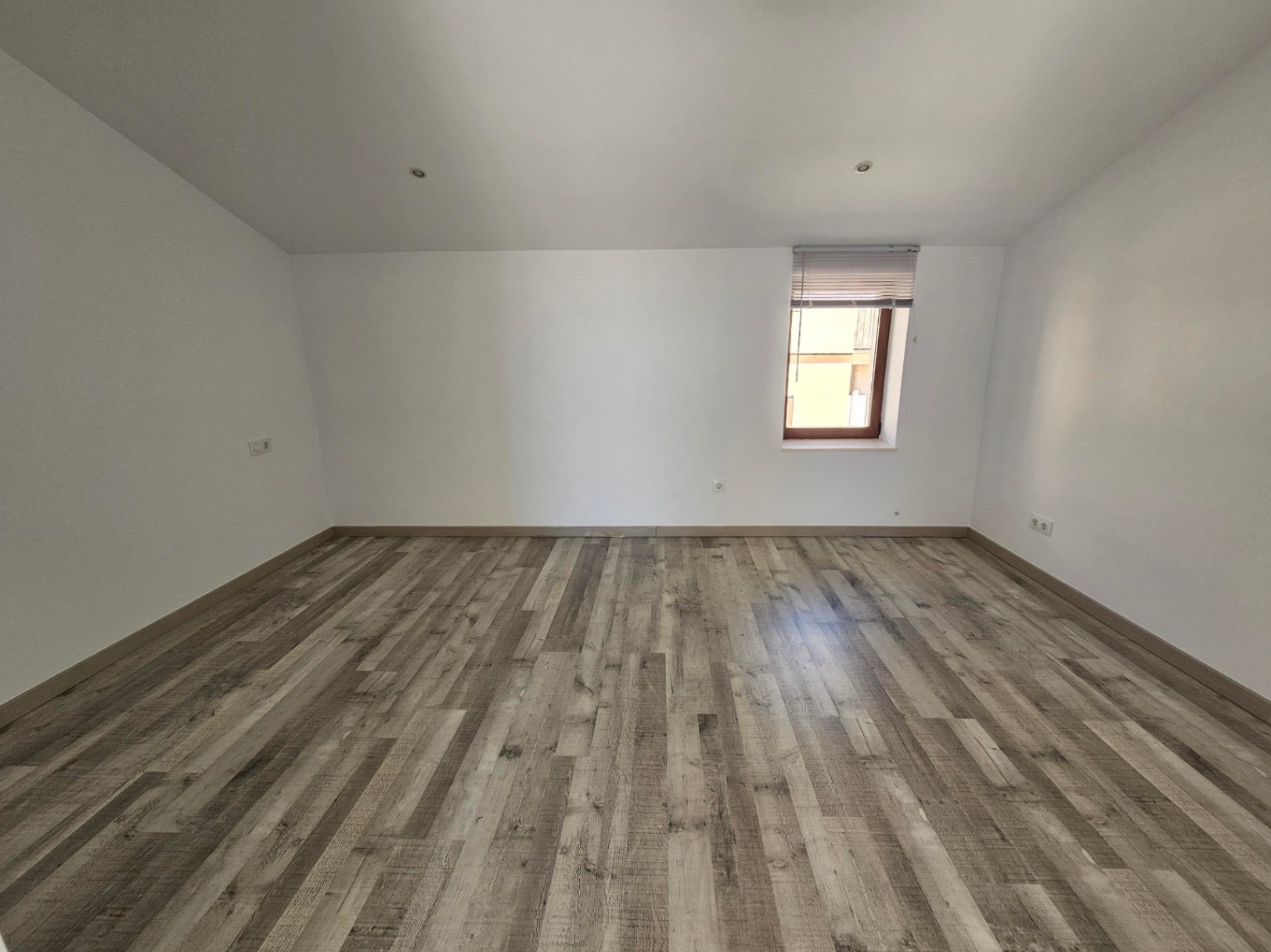 Spacious room featuring grey wood-effect laminate flooring, white walls, and a sloped ceiling with recessed spotlights and a wooden-framed window.
