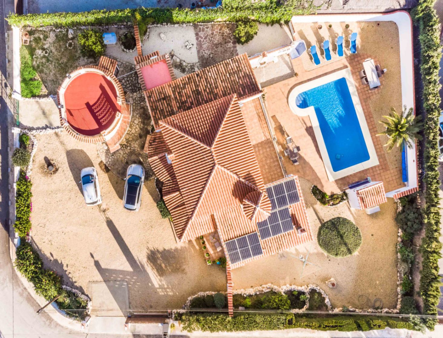 Aerial view showing the villa's terracotta roof with solar panels, the L-shaped swimming pool, sun terrace, and the spacious gravel driveway.