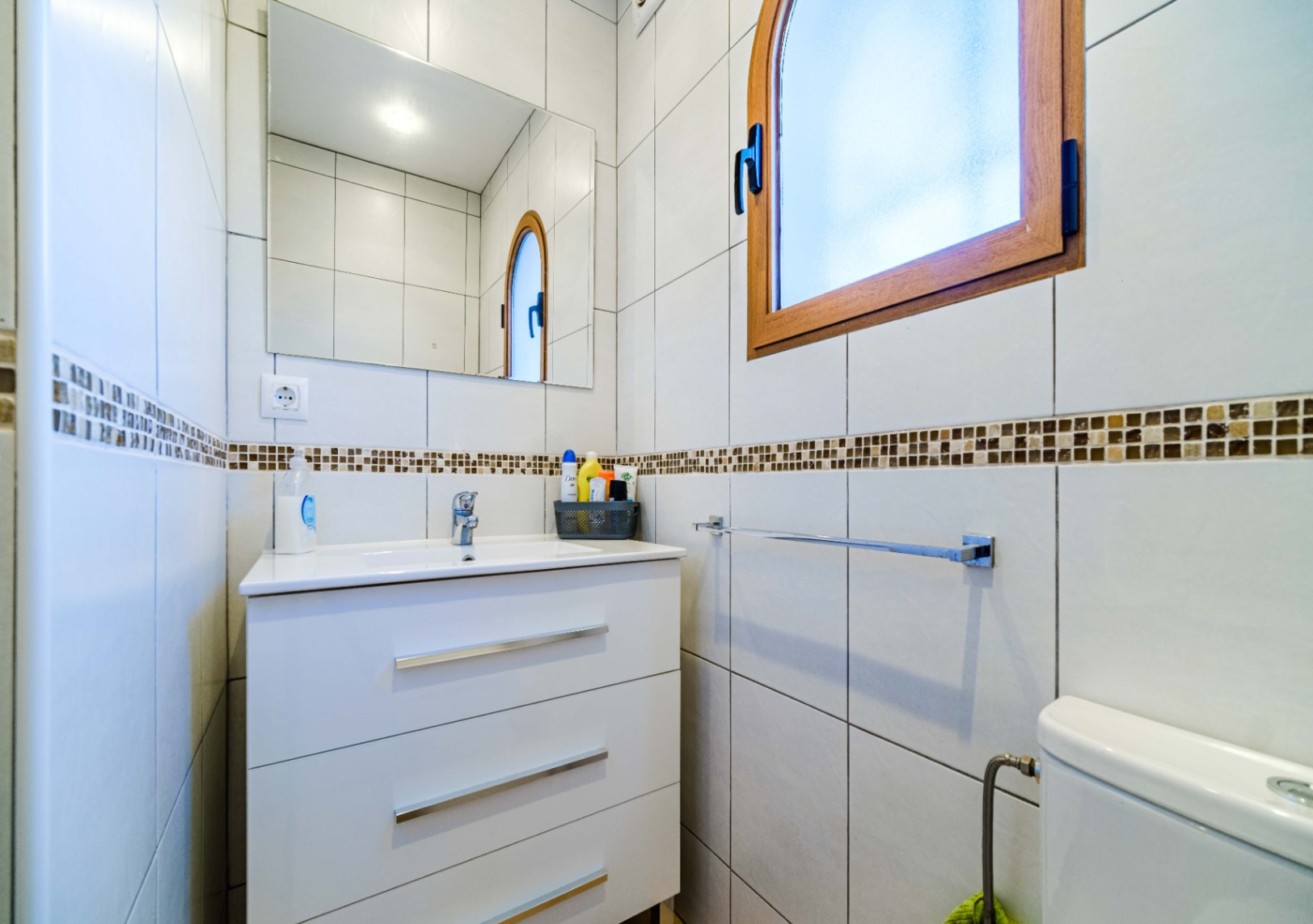 Bathroom featuring a white three-drawer vanity, integrated sink, and rectangular mirror. Includes an arched wooden-framed window and white tiling with a decorative mosaic border.