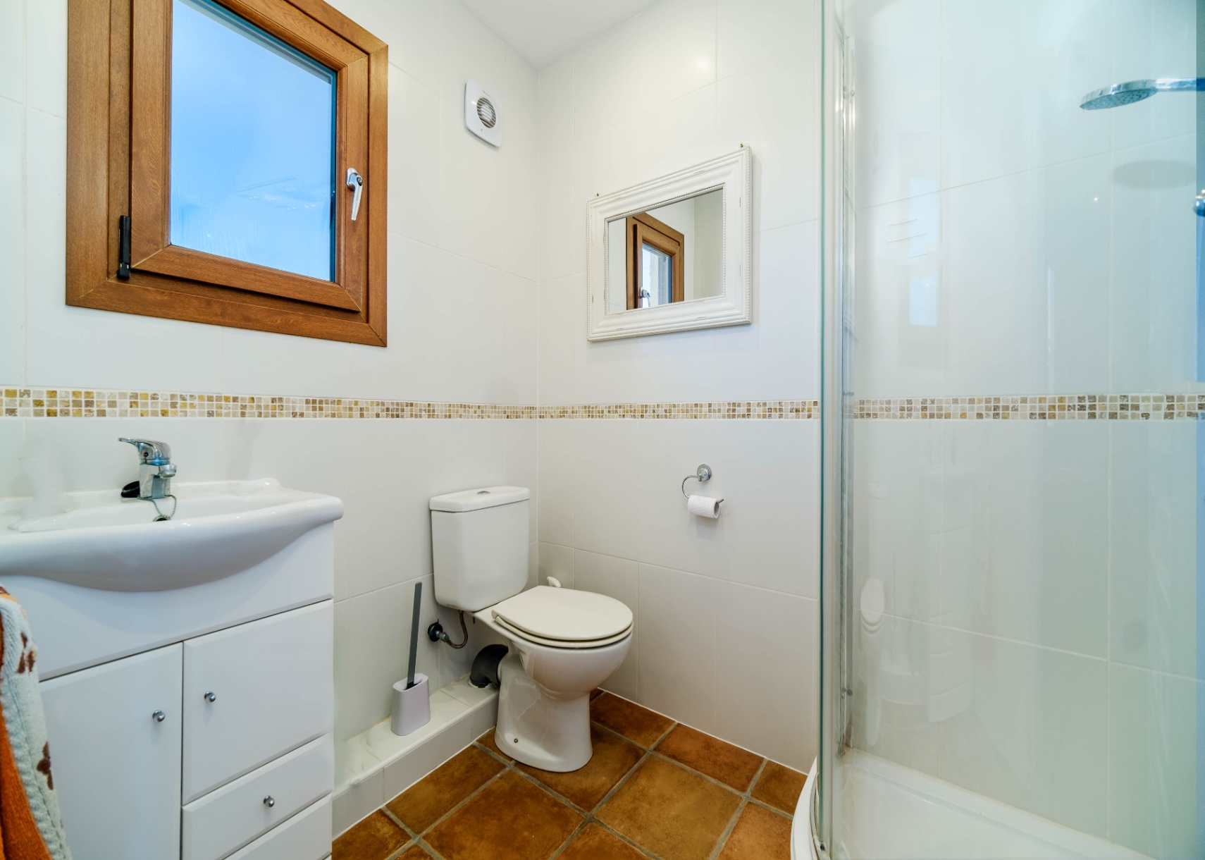 Bathroom featuring white tiled walls with a mosaic border and terracotta flooring. It includes a glass-enclosed shower, a vanity unit, and a wooden-framed window.