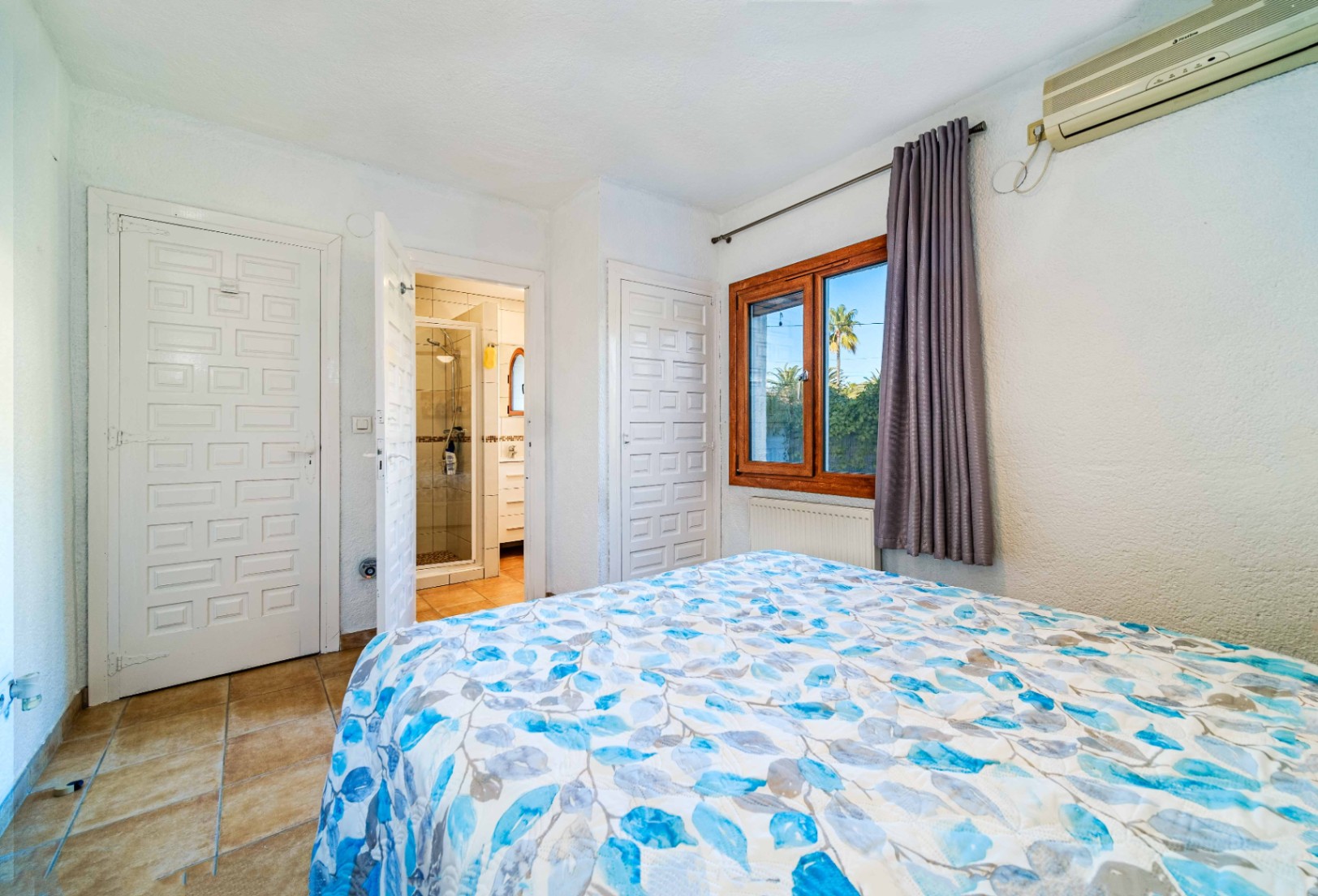Bedroom featuring terracotta tile flooring, an en-suite bathroom with walk-in shower, air conditioning, and a wooden window frame.