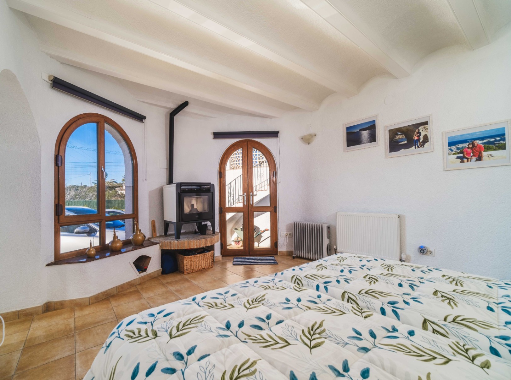 Bedroom featuring white beamed ceilings, a wood-burning stove on a brick plinth, and arched wooden windows that provide ample natural light.