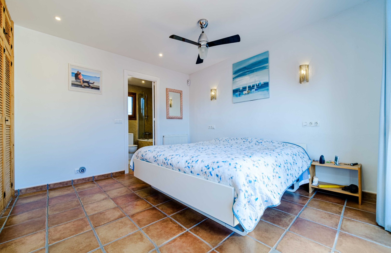 Bright bedroom featuring rustic tile flooring, a ceiling fan, and an en-suite bathroom. Includes built-in wooden wardrobes and integrated wall lighting.