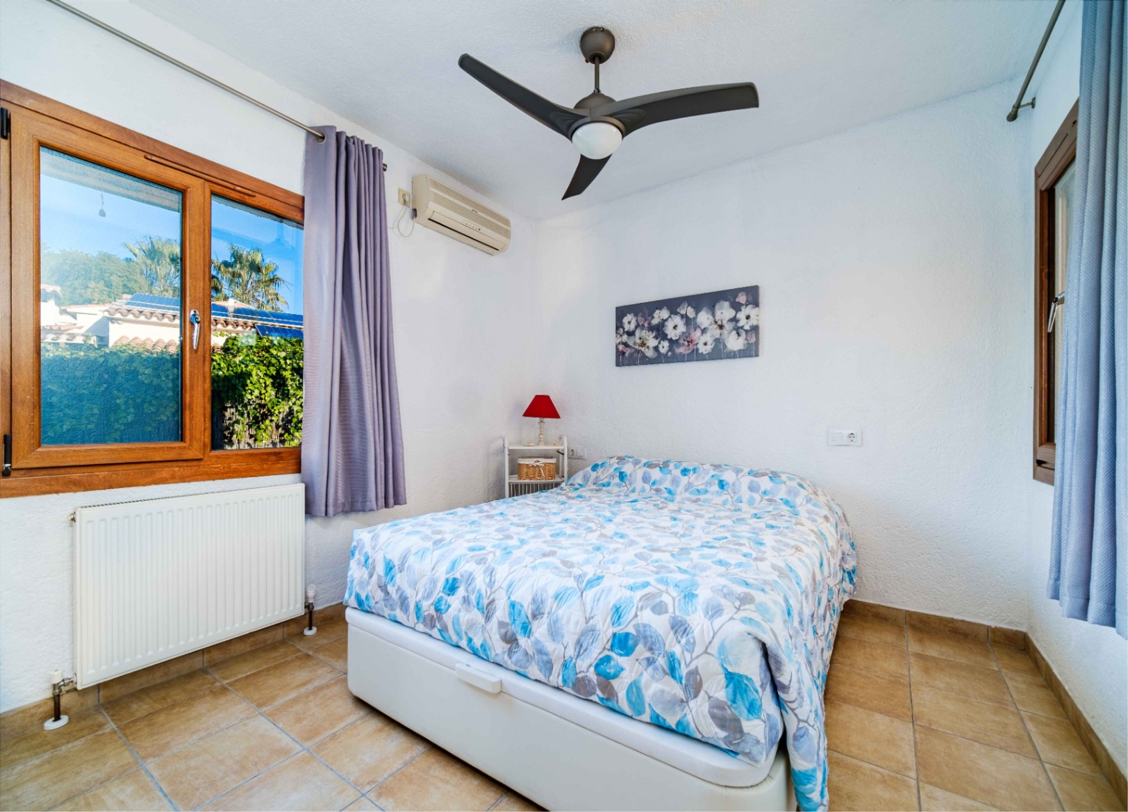 Bright bedroom featuring tiled flooring, a ceiling fan, air conditioning, and wooden-framed windows overlooking the Mediterranean garden.