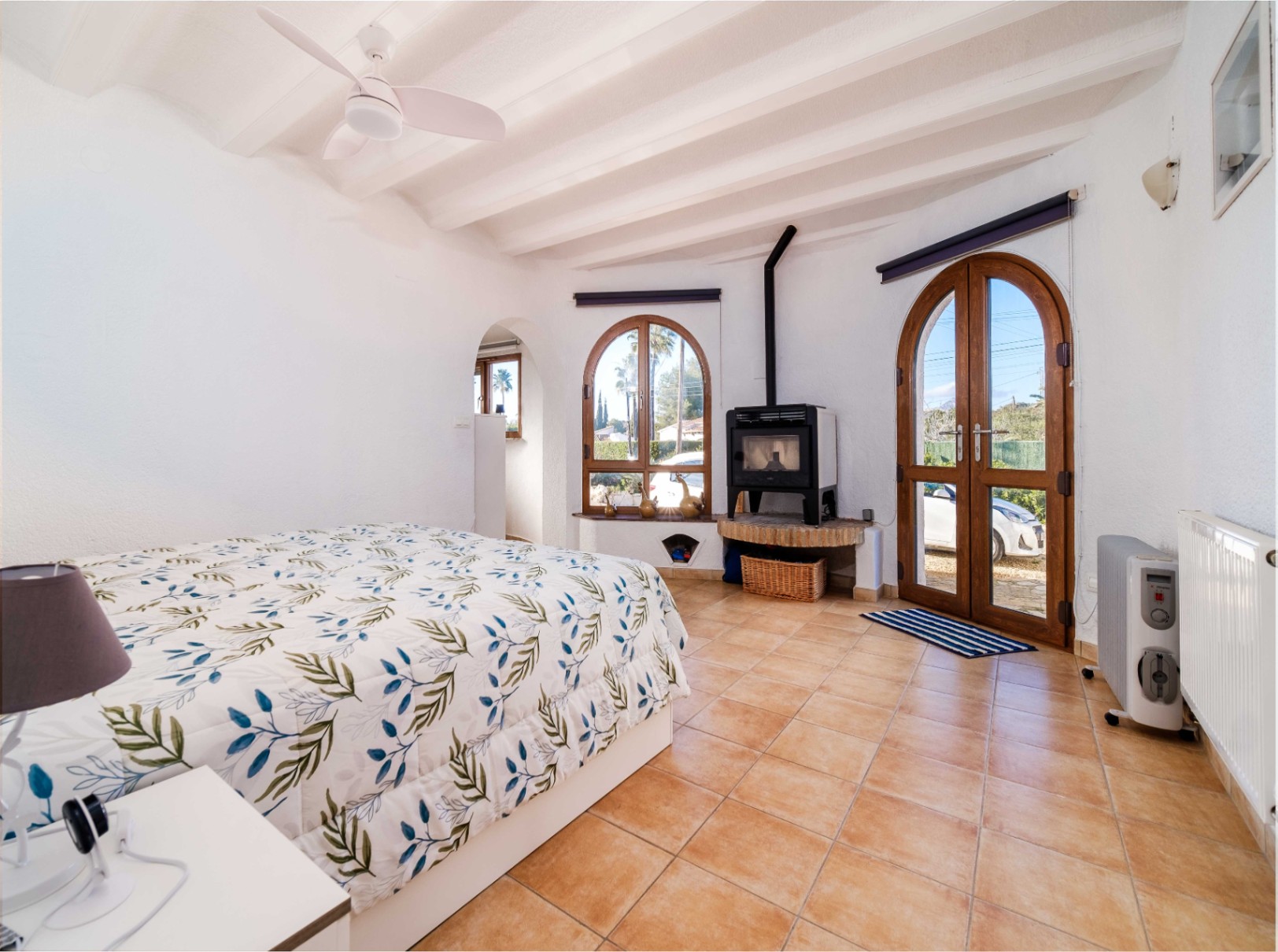 Bright bedroom featuring white beamed ceilings, terracotta tiling, and a wood-burning stove. Arched wooden doors provide direct outdoor access and natural light.