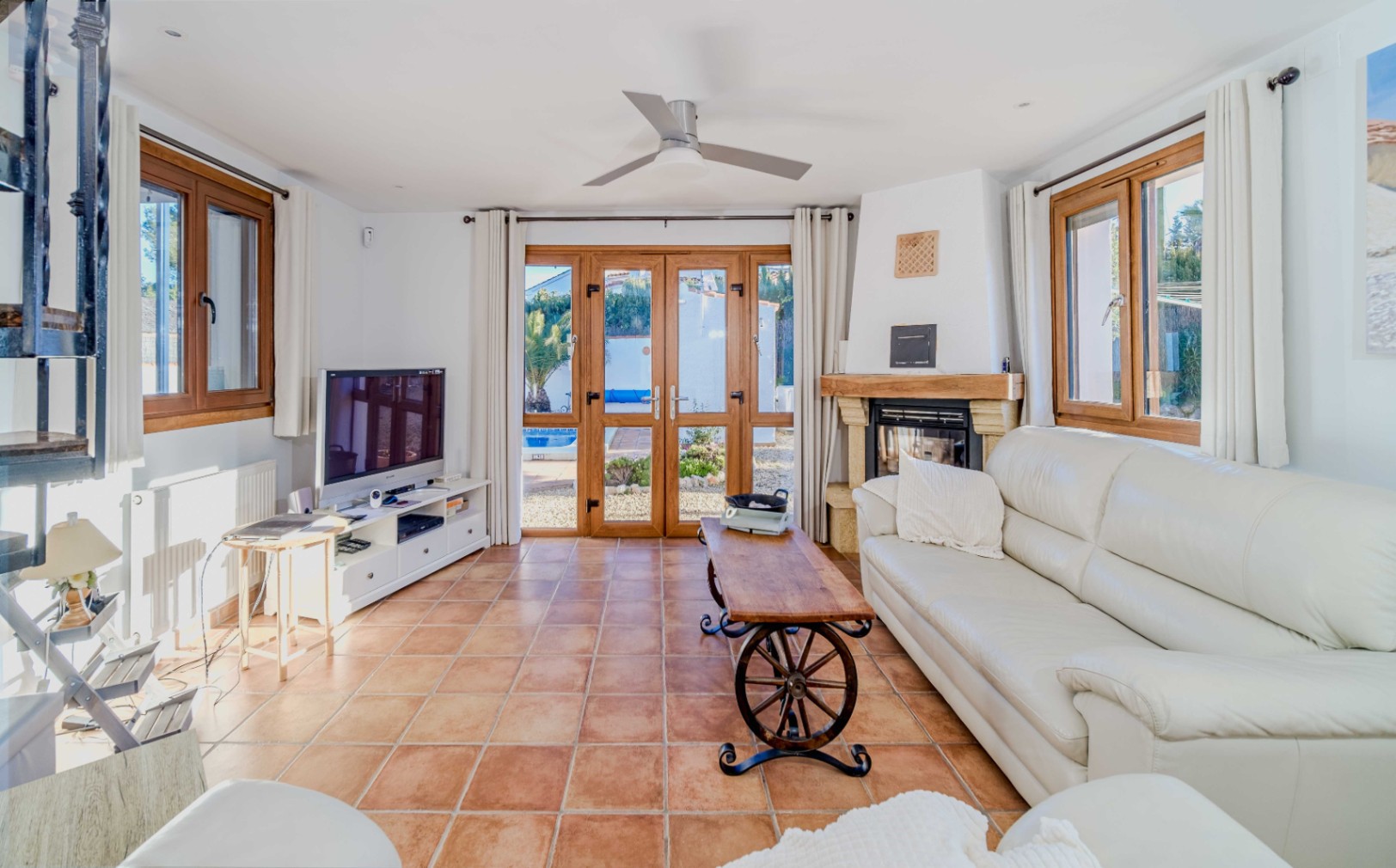 Bright living area featuring terracotta tile flooring, a stone fireplace, and wooden-framed glass doors opening to the pool. Includes a ceiling fan and rustic furnishings.