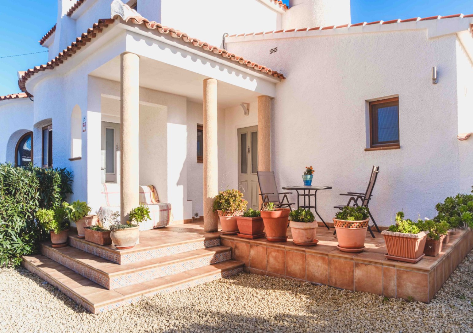 Entrance porch featuring stone columns, terracotta tiling, and decorative step risers. The white facade is accented by potted succulents and a small outdoor seating area.