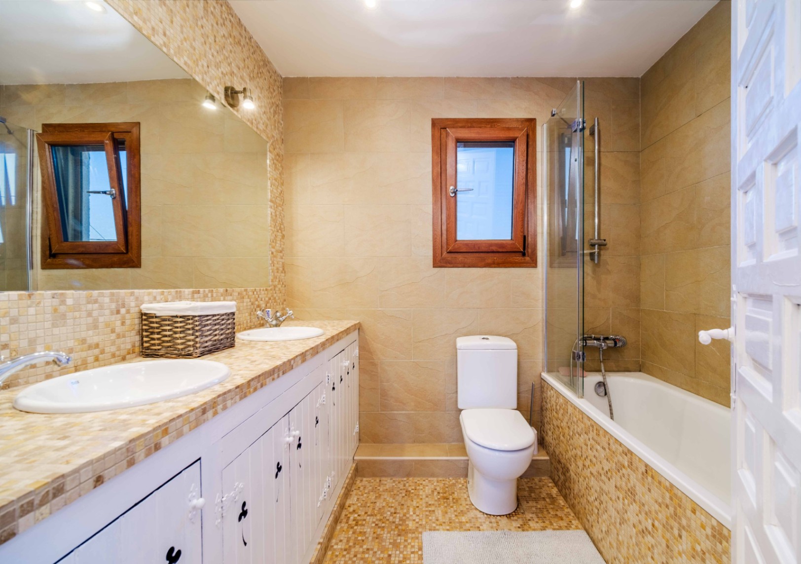 Full bathroom featuring a double vanity with mosaic tiling and white cabinetry. It includes a bathtub with a glass screen, beige wall tiles, and a wooden-framed window.