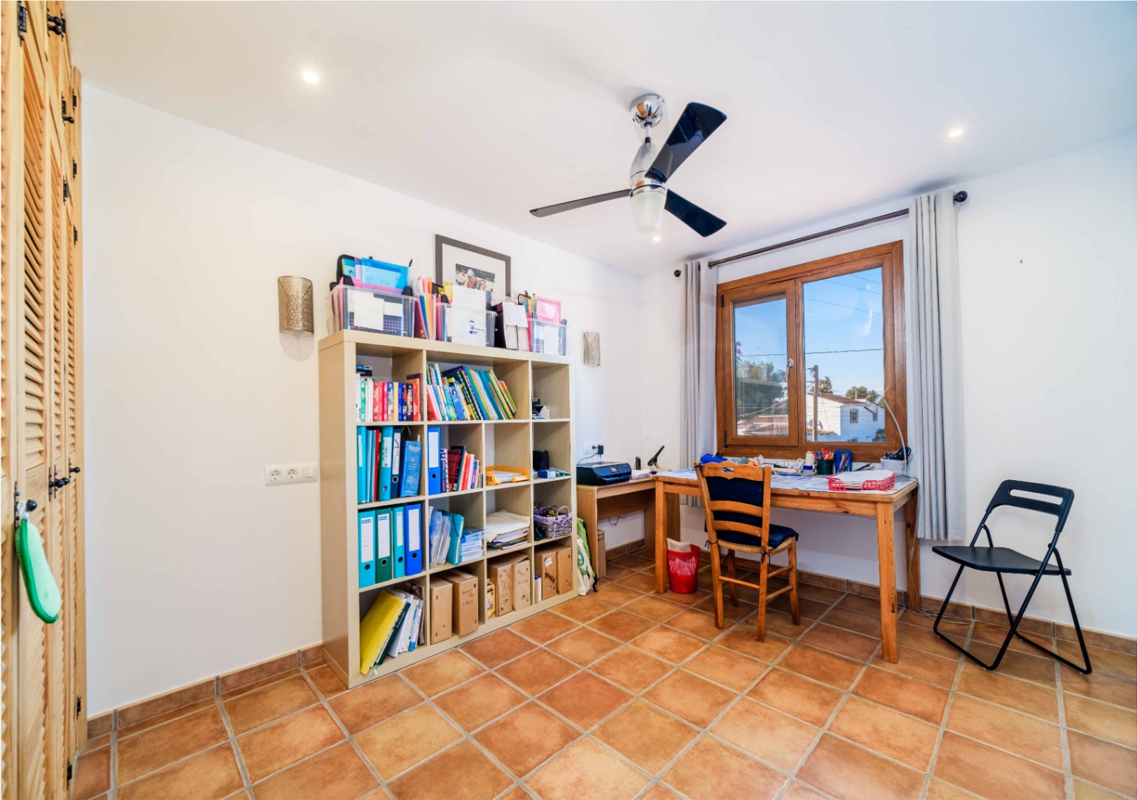 Functional home office featuring terracotta tile flooring, ample modular shelving, a wooden desk by the window, and a modern ceiling fan.