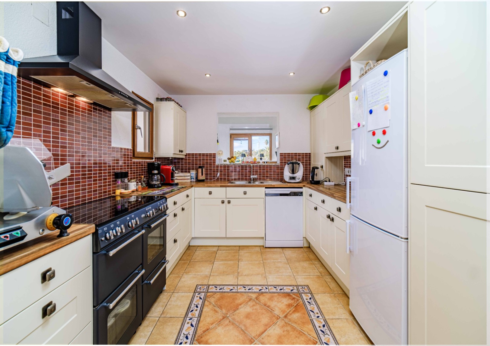 Functional kitchen with white cabinetry, wooden countertops, and a brown tiled backsplash. Features include a large black range cooker and terracotta flooring with a decorative border.