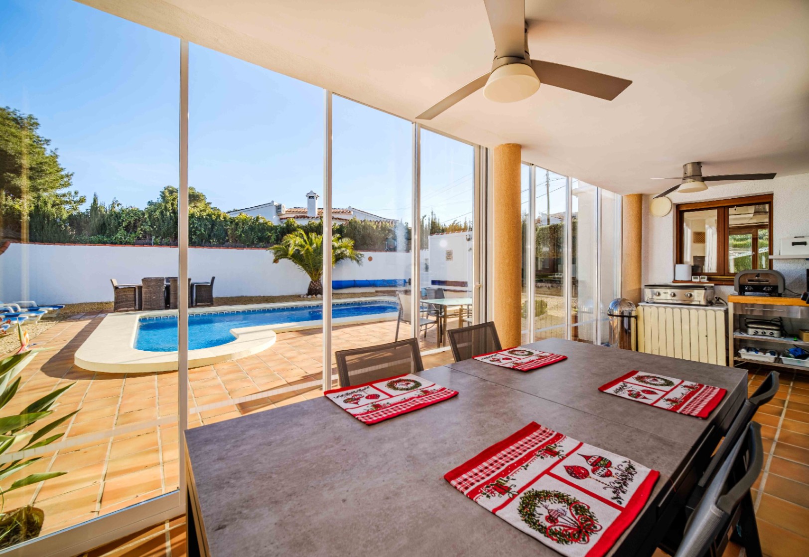 Glass-enclosed terrace featuring a dining area, ceiling fans, and direct views of the private swimming pool and outdoor kitchen.