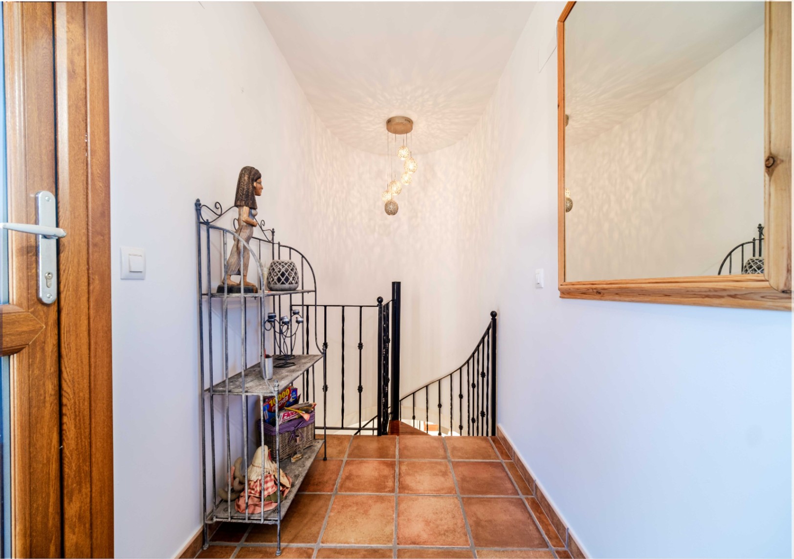Hallway landing featuring terracotta floor tiles, a black wrought iron staircase railing, and a decorative pendant light casting patterns on the walls.