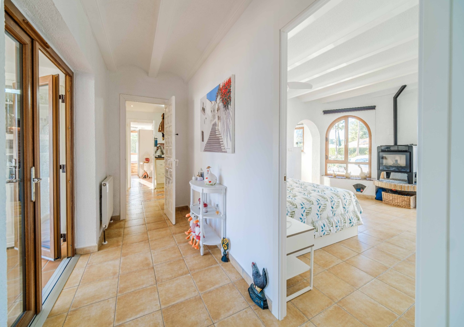 Hallway with terracotta tiling leading to a bright bedroom featuring exposed white beams, an arched window, and a freestanding wood-burning stove.