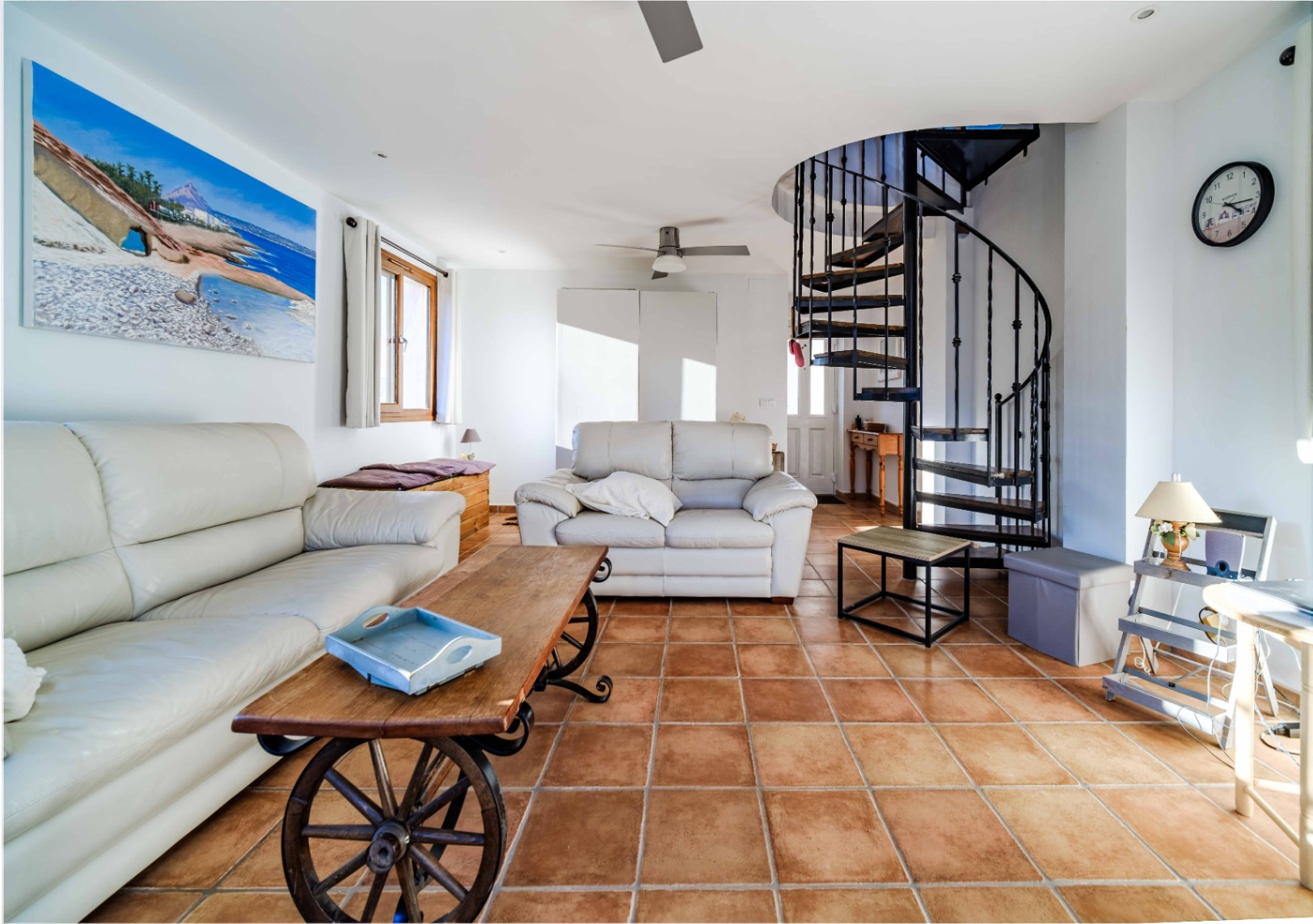 Living area featuring terracotta tile flooring, white leather sofas, and a unique cart-wheel coffee table. A black metal spiral staircase provides access to the upper level.
