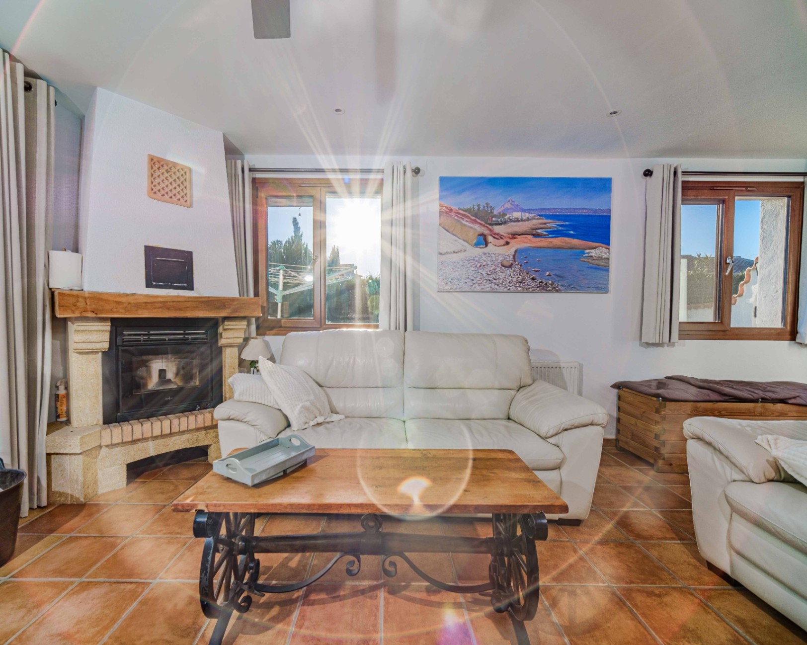 Living room featuring a stone fireplace with a wooden mantel, terracotta tile flooring, and white leather sofas. Natural light enters through wood-framed windows.