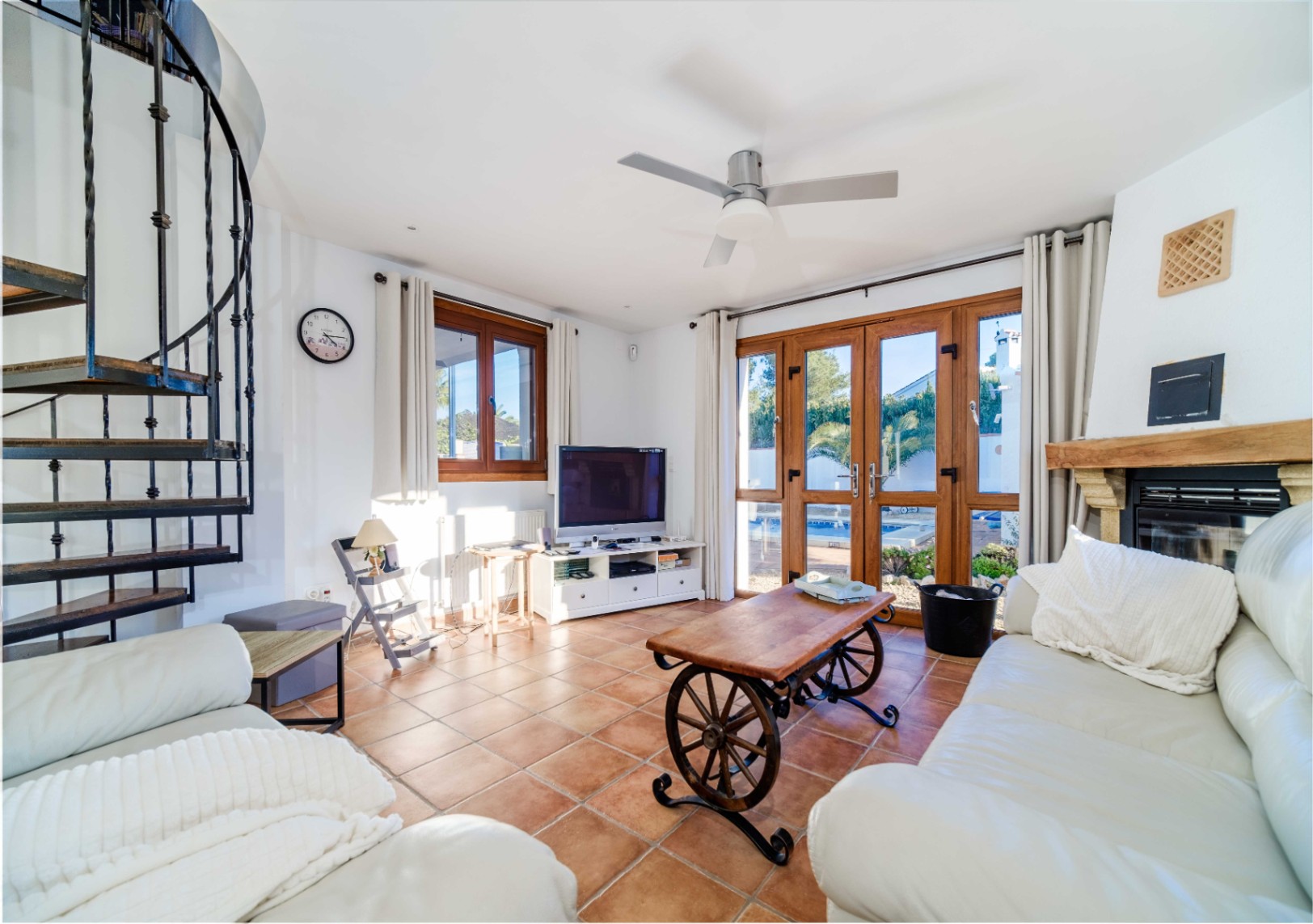 Living room featuring terracotta tiling, a wrought iron spiral staircase, and a fireplace. Large wooden glass doors offer direct views and access to the pool area.