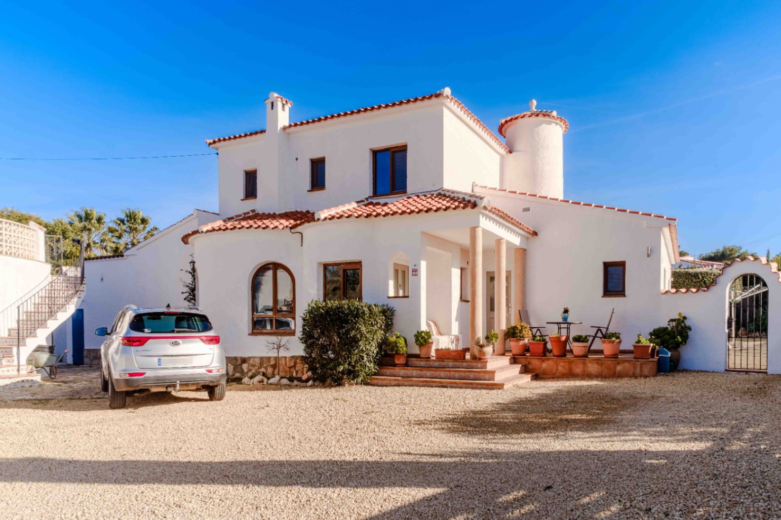 Mediterranean villa exterior featuring white stucco walls, terracotta roof tiles, a columned porch, and a spacious gravel driveway for parking.