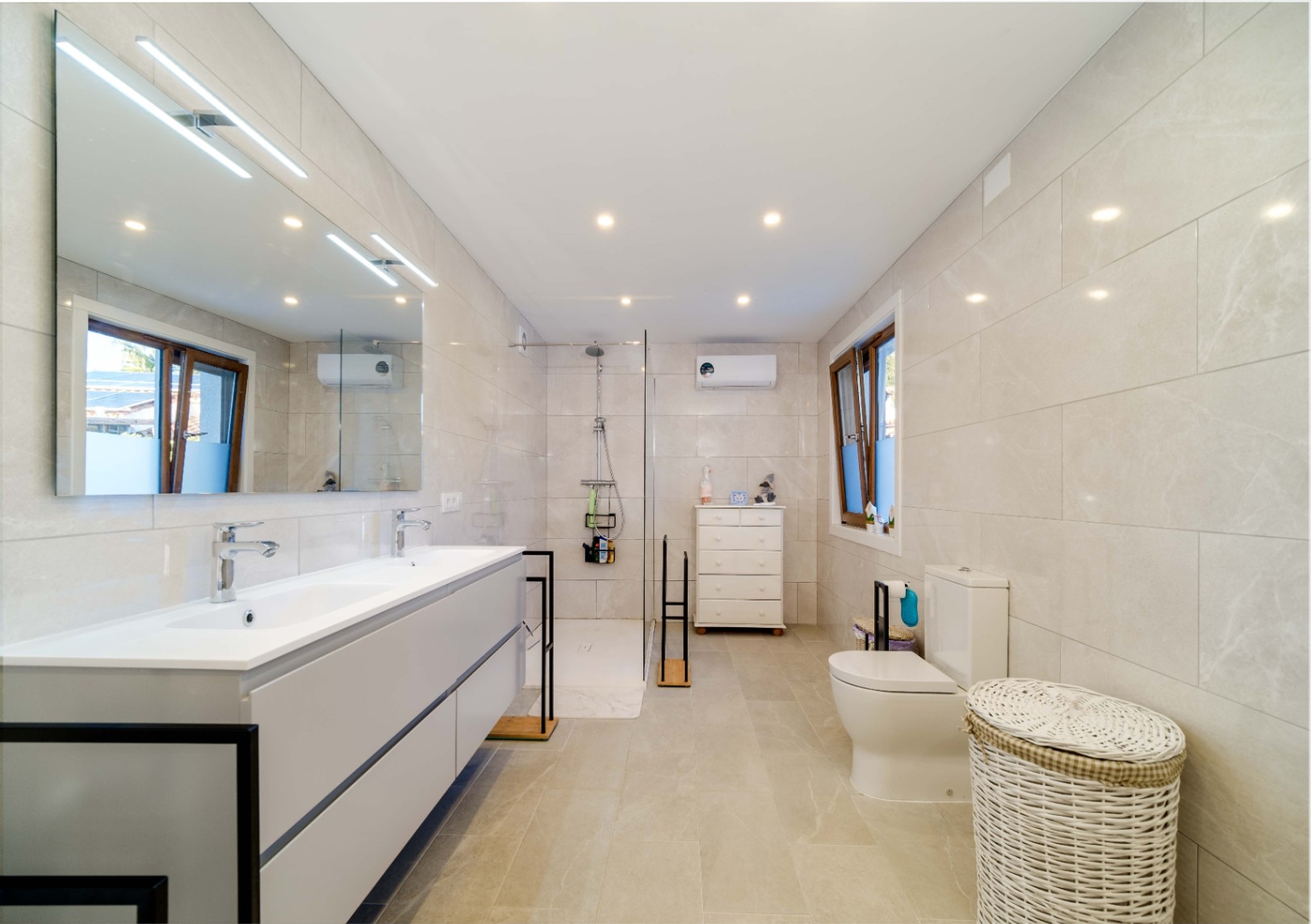 Modern bathroom featuring a double vanity with grey cabinetry, a walk-in shower with glass partition, and neutral porcelain wall tiles with recessed lighting.