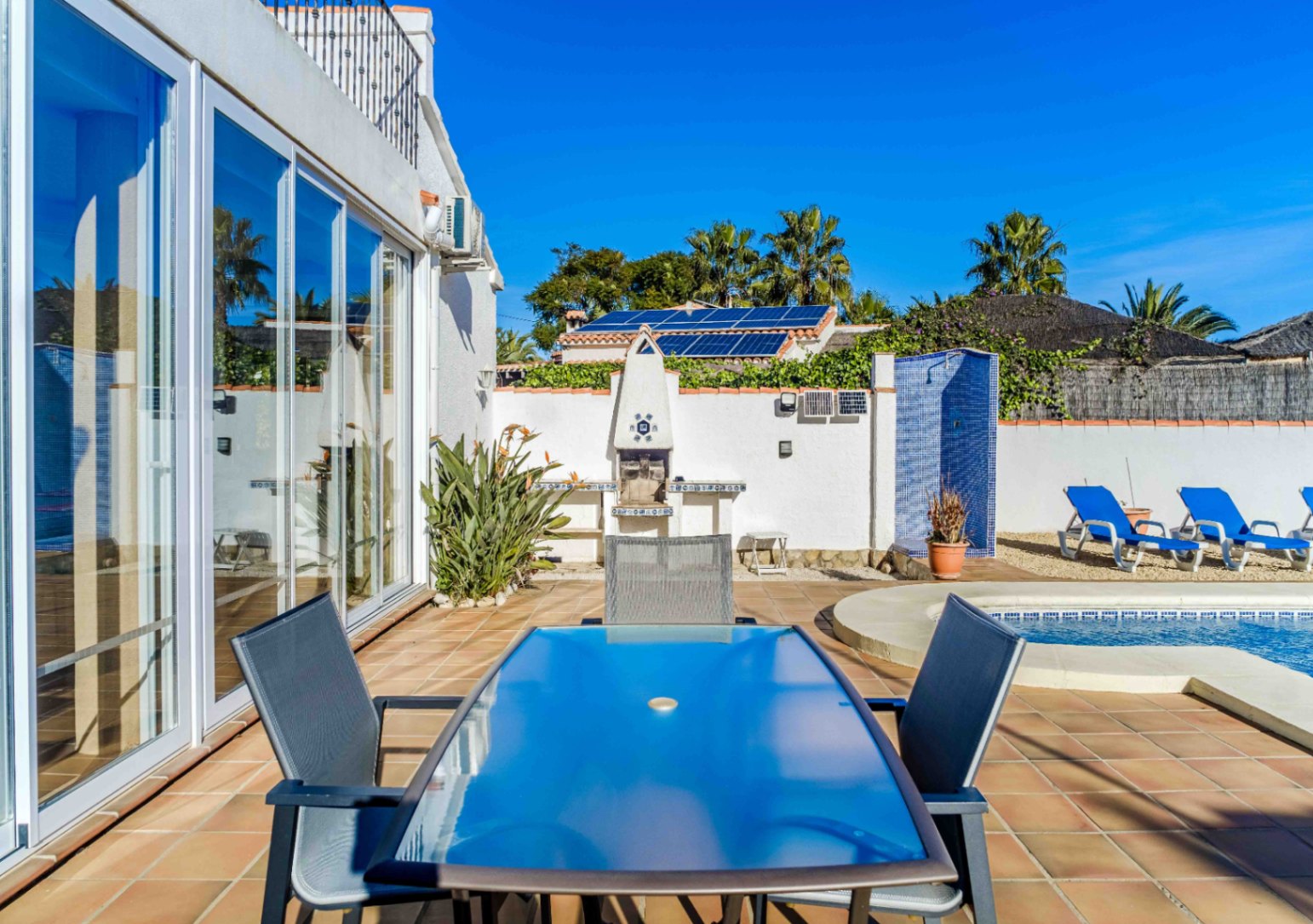 Outdoor terrace featuring a glass-top dining set, built-in barbecue, and blue mosaic shower by the pool. Large glass doors and rooftop solar panels are visible.