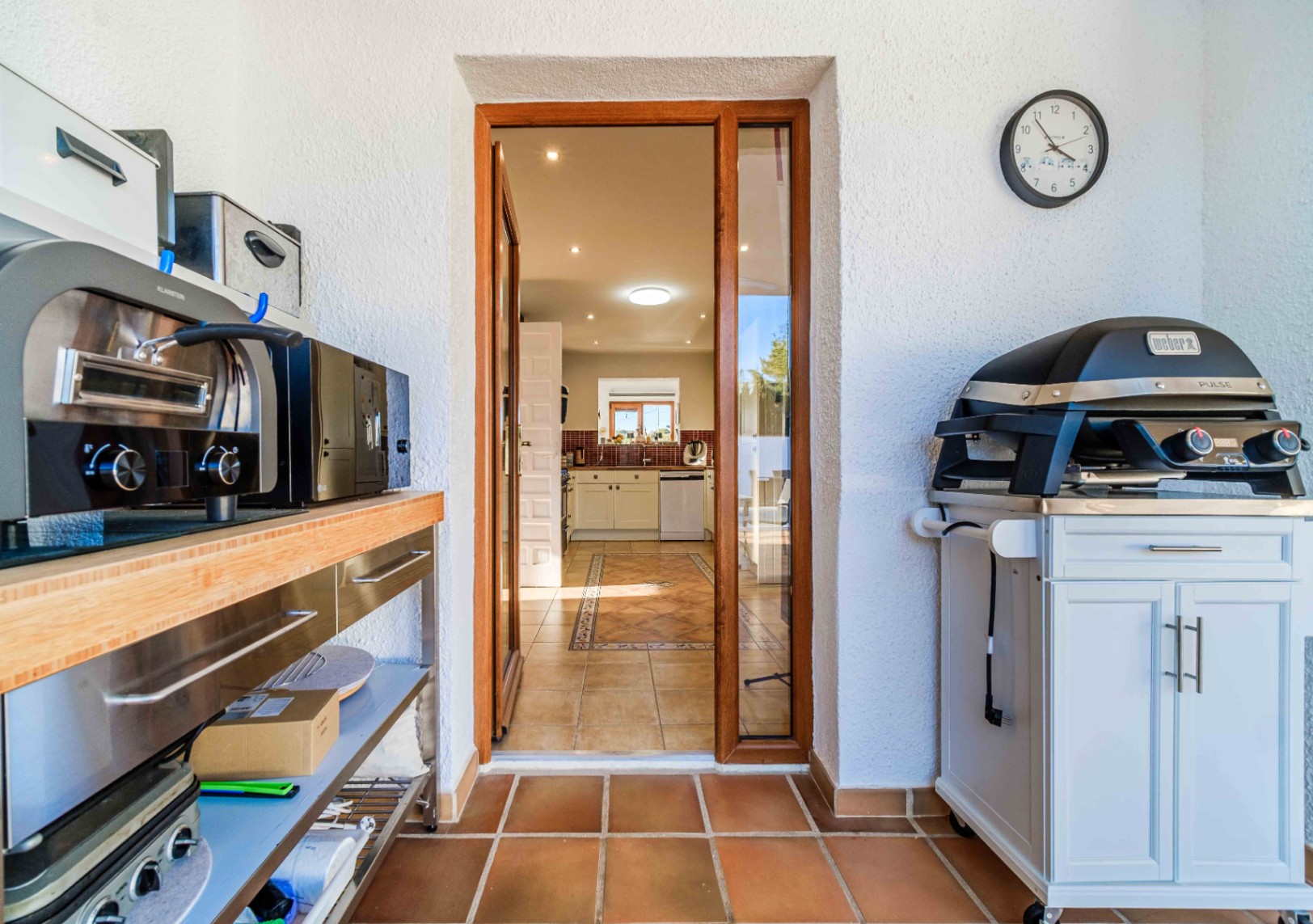 Outdoor utility area featuring a Weber electric grill and stainless steel prep station, leading into the main kitchen through a wood-framed glass door.