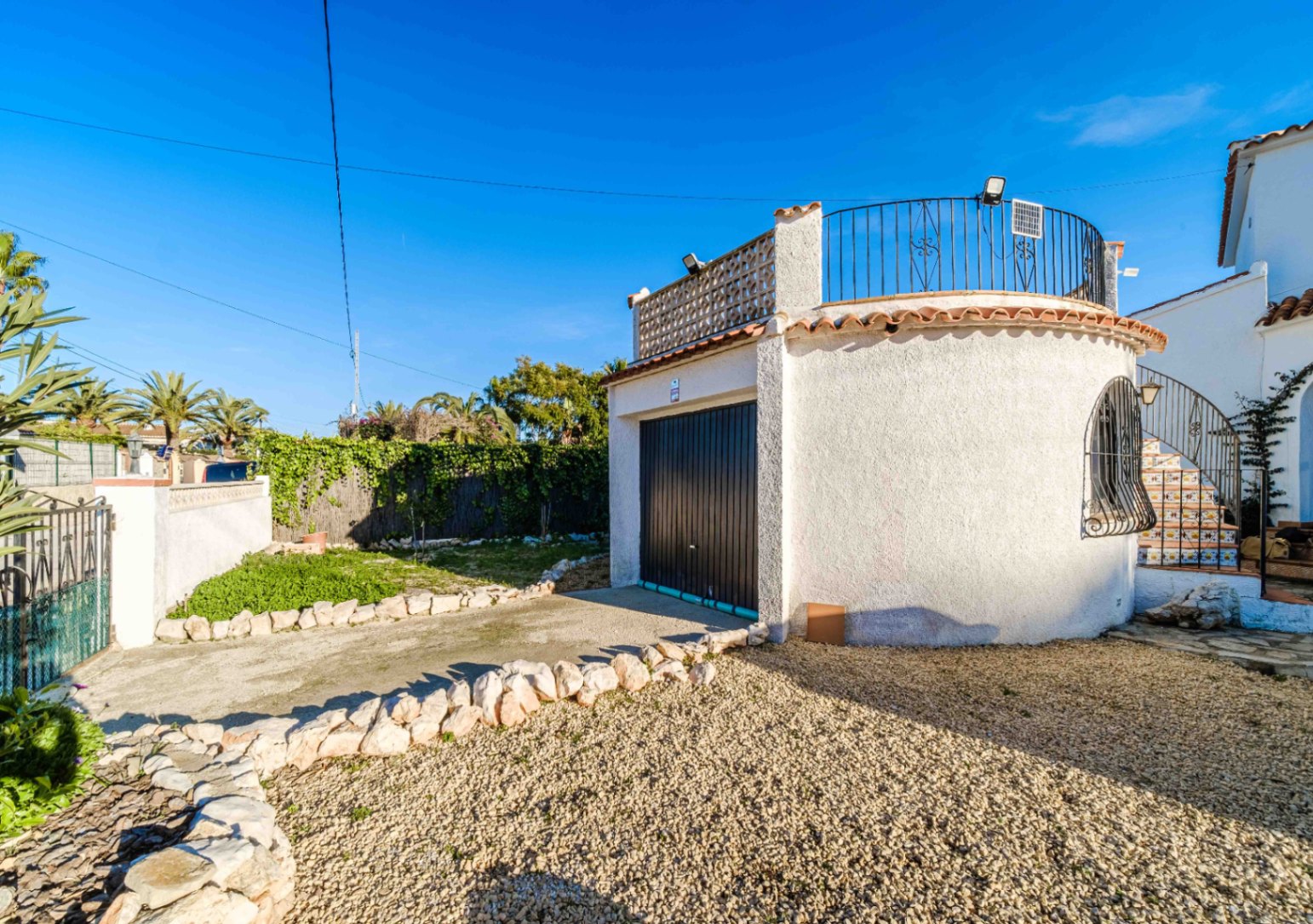 Private garage with a black door and a rooftop solarium featuring wrought-iron railings. The exterior includes a gravel driveway, stone borders, and Mediterranean landscaping.